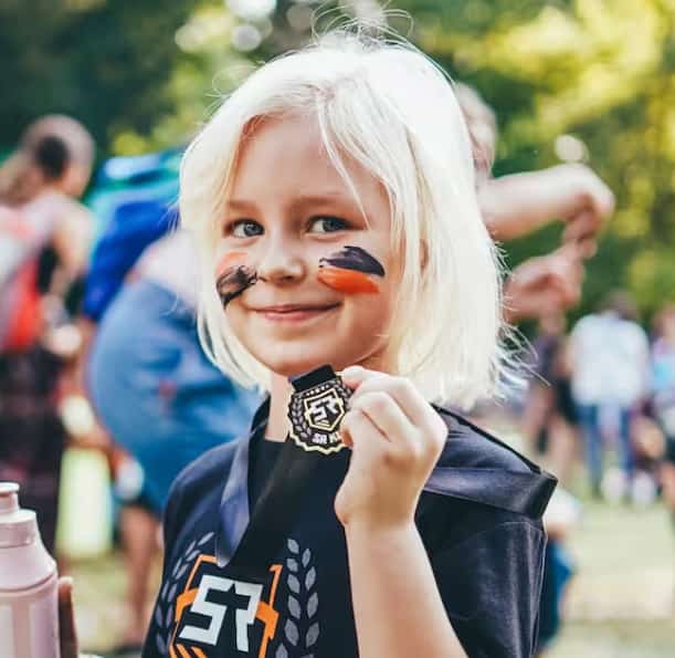 Uśmiechnięta dziewczynka z pomalowaną twarzą trzyma pamiątkowy medal Survival Race Kids po ukończonym biegu z przeszkodami.