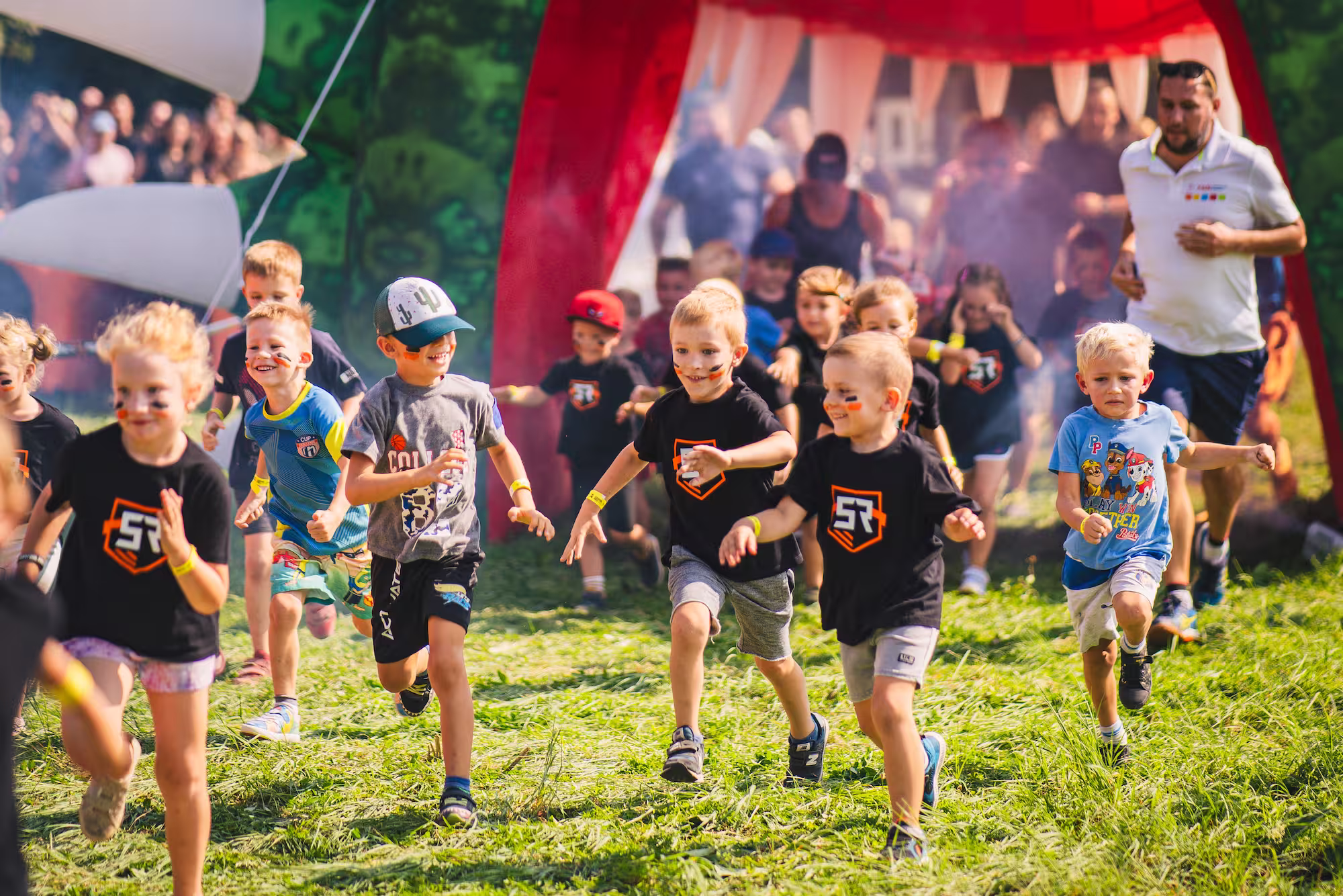 Grupa radosnych dzieci w czarnych koszulkach biegnie przez trawiasty teren podczas sportowego wydarzenia Survival Race Kids.